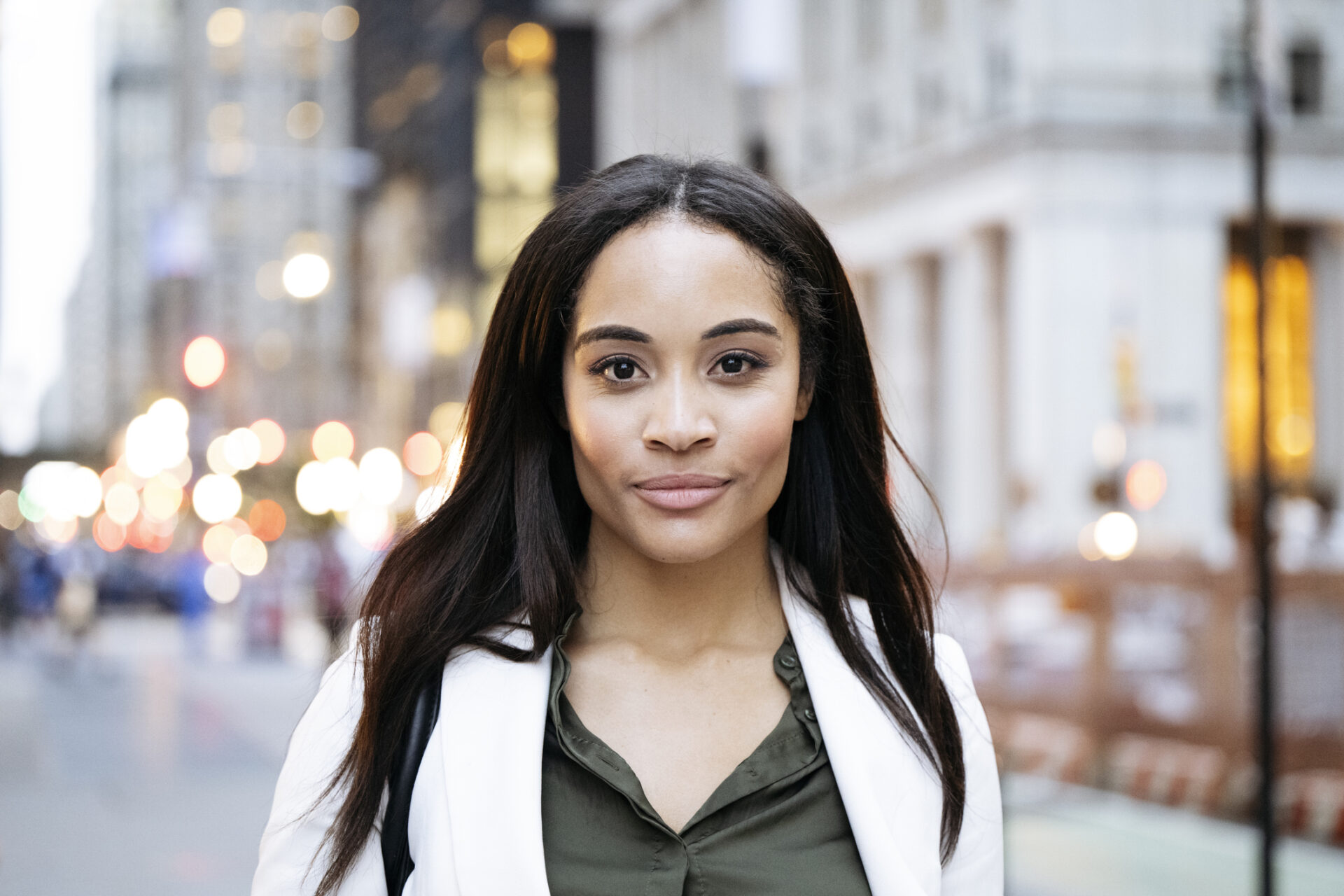 Longhaired professional in late 20s wearing white summer suit, standing outdoors, looking at camera with slight smile, glitter of lights at dusk in background.